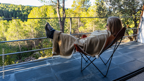 Obraz na płótnie Woman lying on a chair with a blanket wrapped up and enjoying the views of the forest from the terrace. w salonie