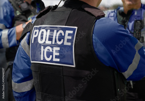 Ice Police Agent - officer of Immigration and Customs Enforcement. Close-up of POLICE ICE marking on the back of a stab proof vest worn by a trio of police officers at the scene of an incident.
