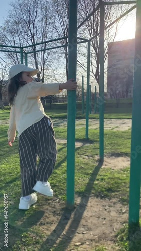 vertical video Young girl wearing a cap, casual shirt, and striped pants balancing on green monkey bars in a sunny playground, fully immersed in the joy of outdoor play and carefree recreation