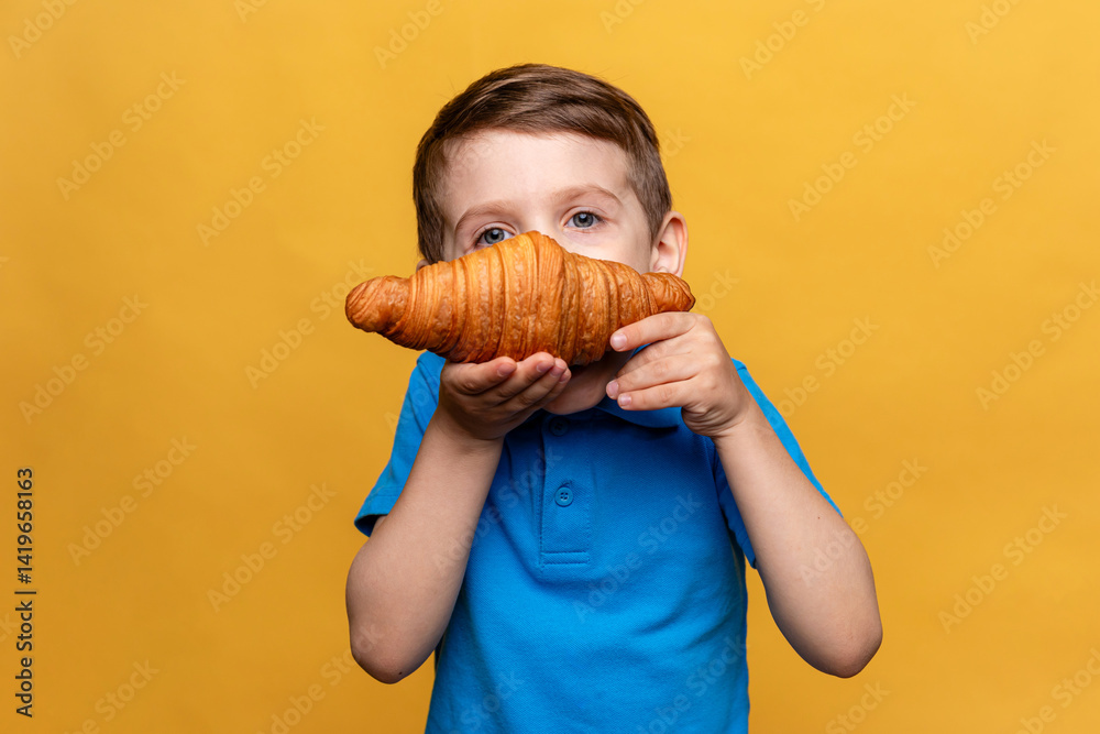 Happy smiling little boy in blue t-shirt eating French croissant on yellow background. Copy space. High quality photo. French breakfast concept.