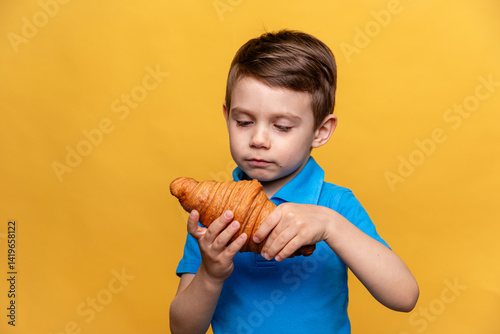 Obraz na płótnie Happy smiling little boy in blue t-shirt eating French croissant on yellow background. Copy space. High quality photo. French breakfast concept. w salonie