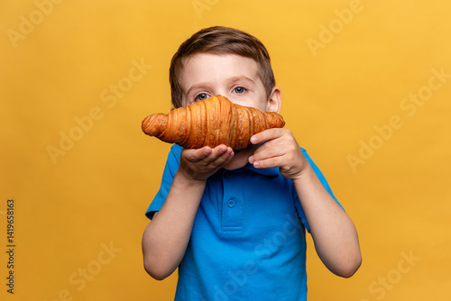 Obraz na płótnie Happy smiling little boy in blue t-shirt eating French croissant on yellow background. Copy space. High quality photo. French breakfast concept. w salonie