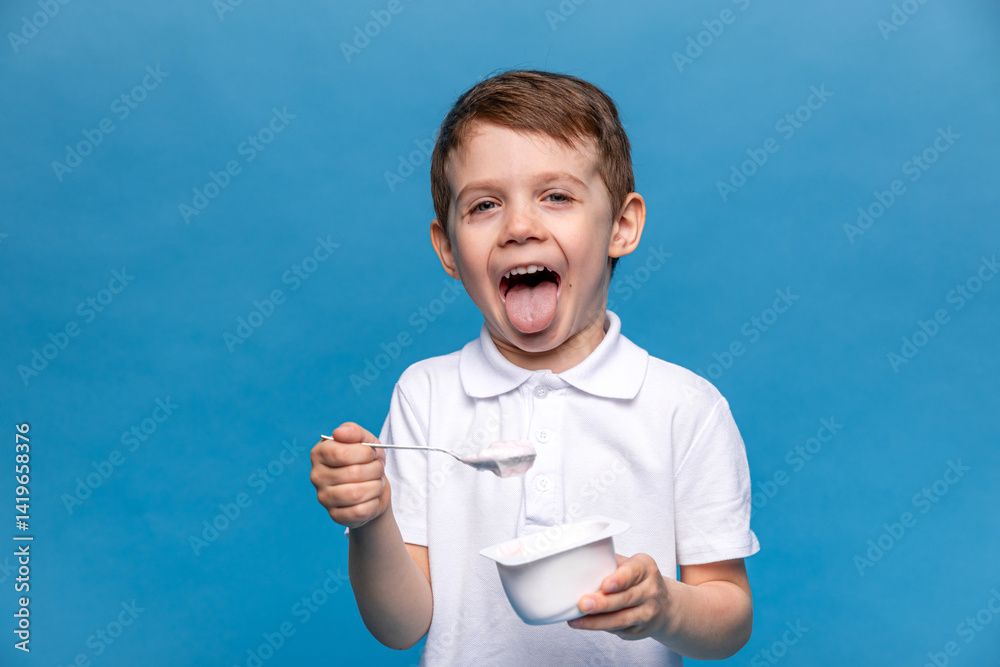 Cute happy laughing little brunette boy, 3-4 years old, with open mouth, in white t-shirt eating tasty yogurt on blue background. Healthy nutrition concept. Copy space. High quality photo