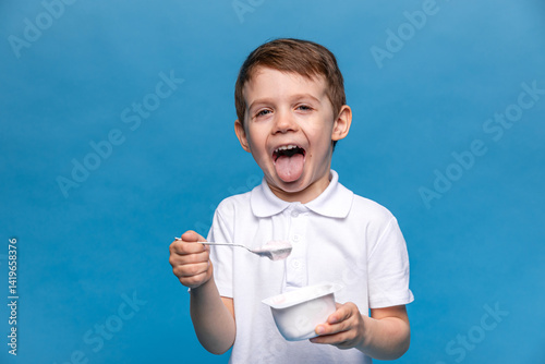 Obraz na płótnie Cute happy laughing little brunette boy, 3-4 years old, with open mouth, in white t-shirt eating tasty yogurt on blue background. Healthy nutrition concept. Copy space. High quality photo w salonie