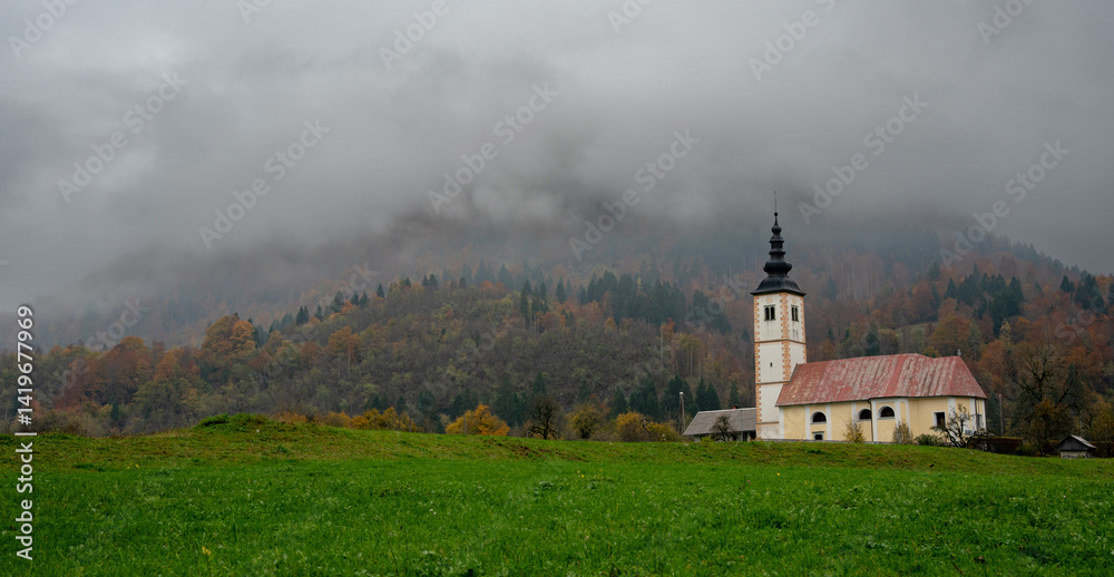 Solitary Cerkev Marijinega church Bitnje standing amidst rolling green hills and misty autumn forest