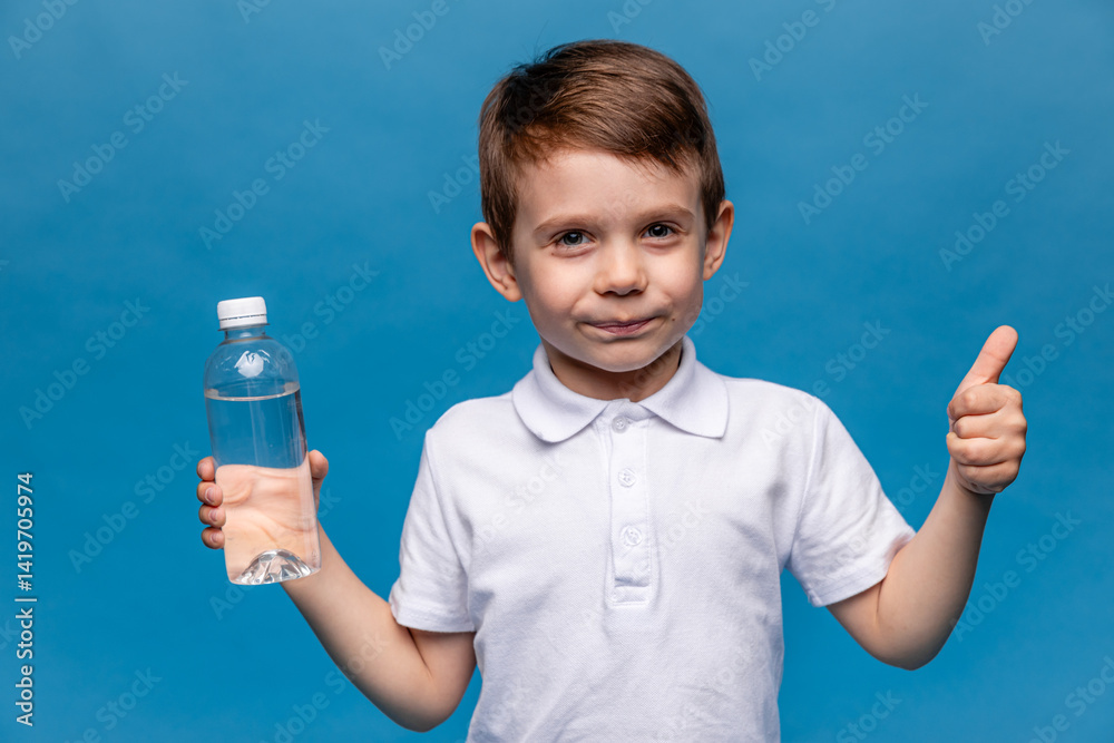 Happy little boy drinks clear water on blue background. The child is holding a plastic bottle of water and showing thumbs up. Children activity and energy, generation Z, body hydration concept
