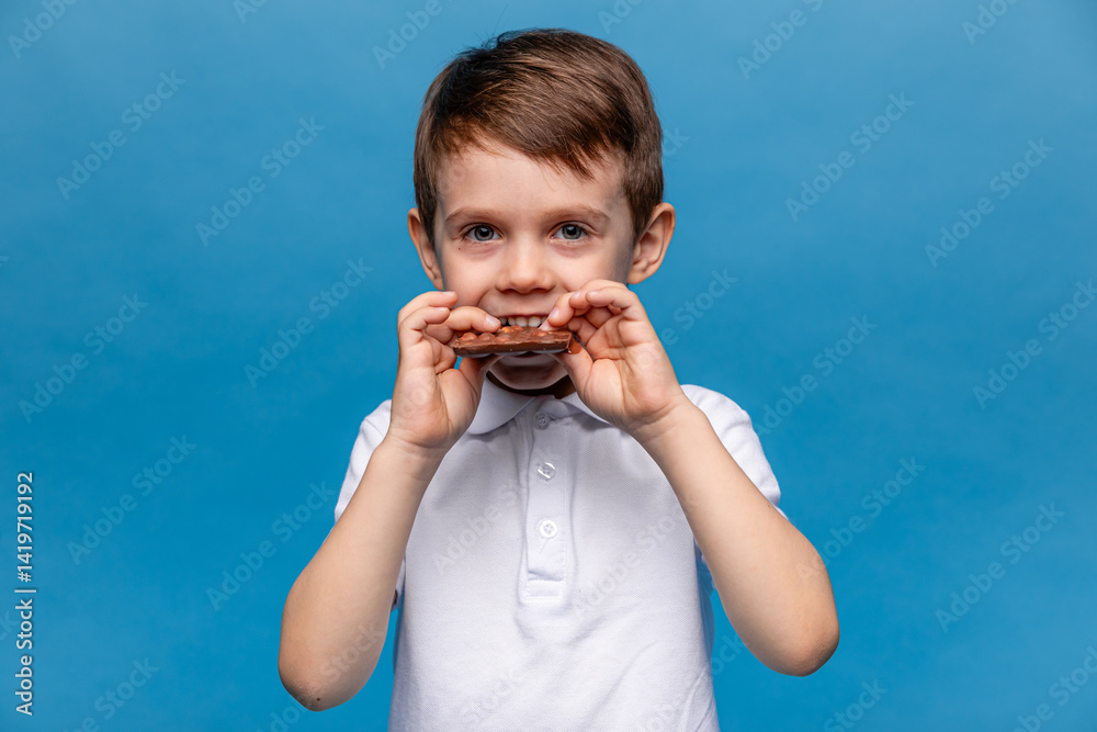 Cute little boy, 3-4 years old, wearing white t-shirt, eating chocolate bars with nuts on blue background, closeup. The concept of children's happiness and love for sweets. High quality photo