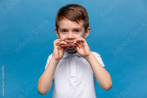 Obraz na płótnie Cute little boy, 3-4 years old, wearing white t-shirt, eating chocolate bars with nuts on blue background, closeup. The concept of children's happiness and love for sweets. High quality photo w salonie