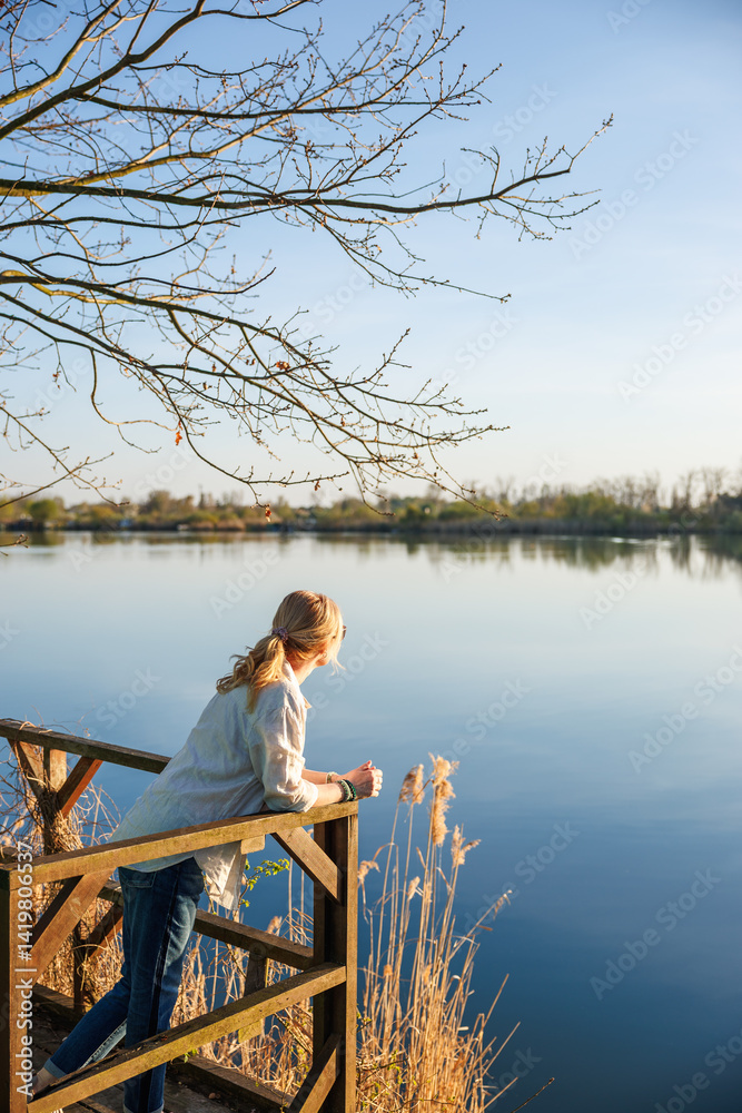 Woman standing on wooden terrace, looking at calm lake and enjoying quiet moment in nature. Contemplation outdoors