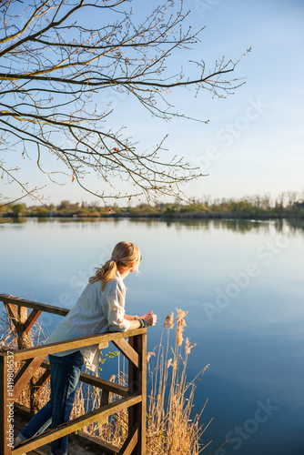 Obraz na płótnie Woman standing on wooden terrace, looking at calm lake and enjoying quiet moment in nature. Contemplation outdoors w salonie