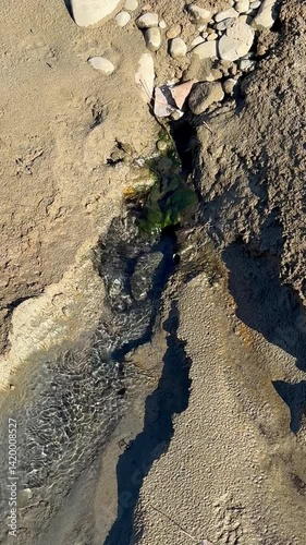 A steady stream of water flows over dry, cracked earth and textured mud, forming natural patterns with reflections and erosion marks. Ideal for nature themes, backgrounds, and geology visuals