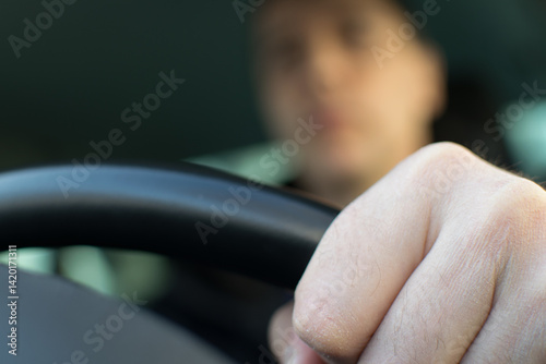 The driver's left hand holds the steering wheel against the background of a blurred face in the car interior