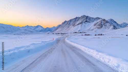 Winter road winding through snowy mountains at sunrise.  A scenic, empty road, blanketed in pristine snow, stretches into the distance, leading toward a mountain range. The peaks are capped with snow,