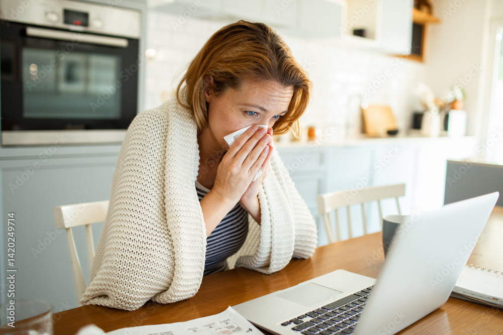 Sick woman blowing nose in tissue at kitchen table with laptop