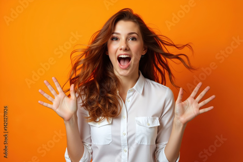 Obraz na płótnie A woman with long brown hair, wearing a white shirt, looking surprised with her hands raised, against an orange background. w salonie