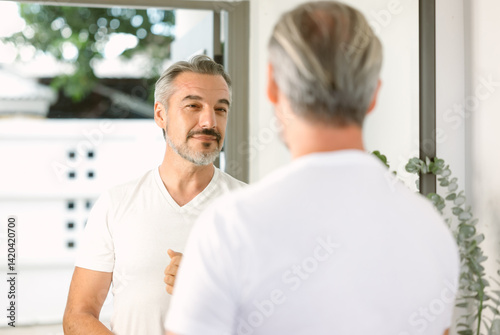 Handsome middle-aged Caucasian man with beard looks in the mirror healthy skin at home. Middle aged man taking care of himself. Morning routine. Receding hairline, haircare, beauty concept.