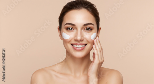 Portrait of a cheerful woman gently applying a skincare cream to her face, showcasing healthy skin and self-care routines, captured on a plain beige background, emphasizing beauty and wellness.

