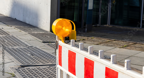 A construction barrier with a bright yellow warning light on a city sidewalk, signaling ongoing work, showcasing urban infrastructure in a modern setting.