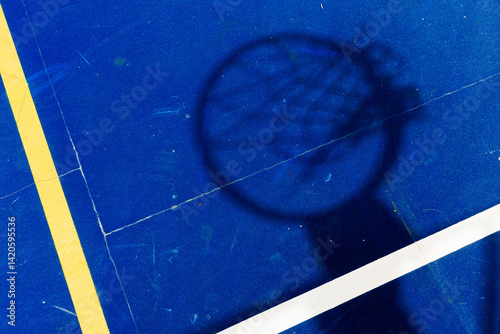 Fototapeta Shadow of a basketball hoop cast on a vivid blue court with yellow and white lines, viewed from above for graphic impact