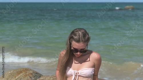Beautiful woman in a pink bikini walking and posing gracefully on a rocky beach. Morning sunlight, clear blue sky, and turquoise water create a serene summer scene