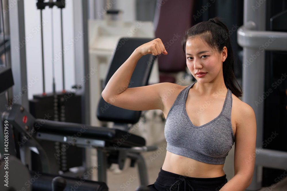 Confident young woman in sportswear flexing arm muscles in modern fitness center.
