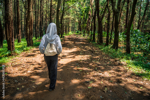 Back view of a woman wearing a hijab carrying a small bag walking into a pine forest while enjoying the fresh air, a woman walking into a pine forest.