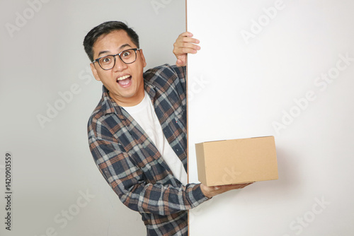 Φωτογραφία Excited Asian delivery man or courier peeks out from behind a white wall with copy space, holding a cardboard box, isolated on a white background