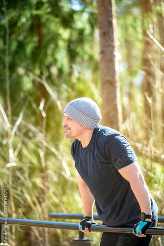 Fit man training outdoors on parallel bars in a forest setting. Wearing a beanie and gloves, he showcases strength and determination. Fitness, wellness, outdoor sports and healthy lifestyle concept