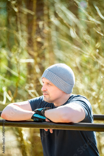 Athletic man resting after outdoor workout, leaning on parallel bars in forest gym. Wearing a beanie and gloves, he looks thoughtful and focused. Fitness, motivation and wellness concept