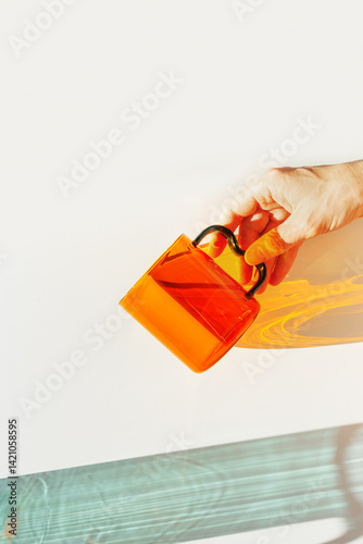 Stylish still-life with orange transparent glass mug in women hand shining orange cup. Sunlight dynamic yellow shadows, reflections on white, minimal aesthetic design, modern kitchen, vibrant shade