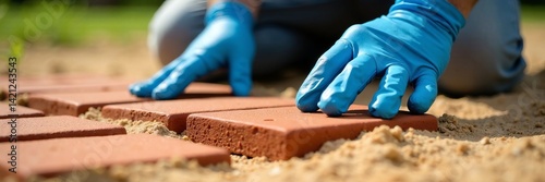 A close-up of hands wearing gloves laying bricks on a sandy surface for landscaping.