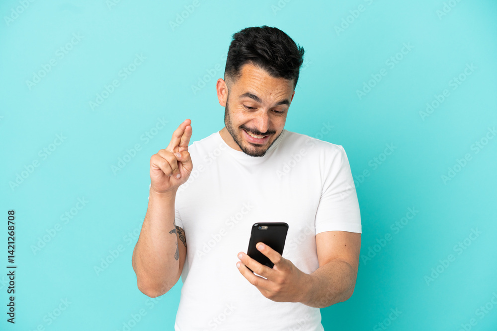 Young caucasian man isolated on blue background using mobile phone with fingers crossing Young caucasian man isolated on blue background using mobile phone with fingers crossing