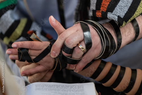 With the help of another adult, a bar mitzvah boy lays the black leather straps of his tefillin around his arm, hand and fingers in preparation for Jewish morning prayers.