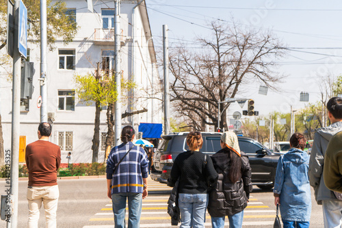 A group of people stands patiently at a crosswalk in an urban setting, waiting for the traffic light to change. The bright sunlight casts shadows on the pavement and cars are stopped in the background