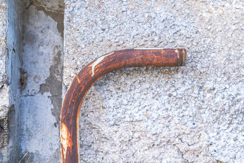 This image shows a bent, rusty metal pipe leaning against a rough-textured concrete wall. The photograph emphasizes the contrast between the corroded reddish-brown metal and the gritty cement surface