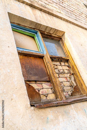 This image captures a decaying window in a crumbling building facade. The wooden frame is peeling, the glass is foggy and cracked, and the lower section has been boarded with rusting metal sheets