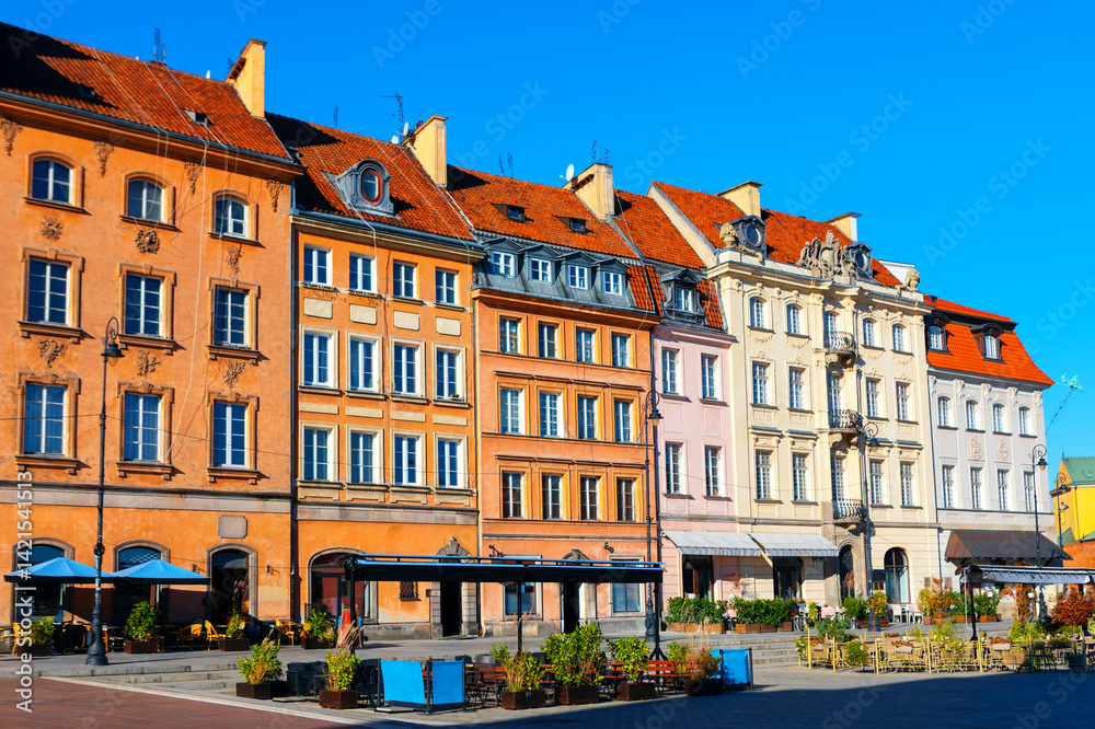 Colorful historic buildings line a sunny square in Warsaw Old Town, showcasing charming architecture and inviting cafes. Clear blue sky adds to the vibrant, picturesque atmosphere