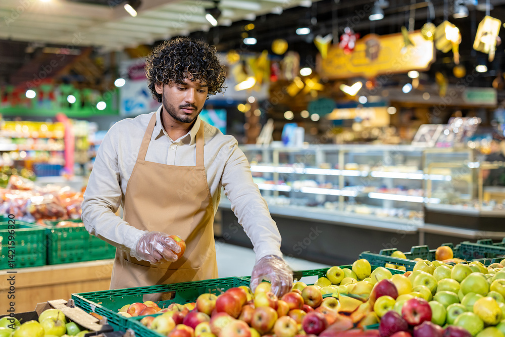 A grocery worker wearing an apron and gloves carefully selects apples from a fresh produce display, checking for quality.