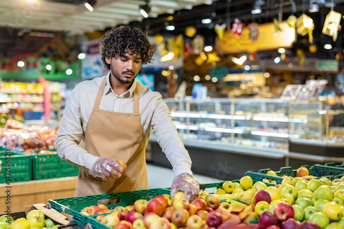 A grocery worker wearing an apron and gloves carefully selects apples from a fresh produce display, checking for quality.