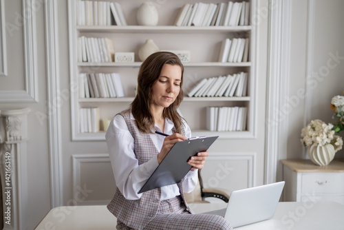 a woman in a business suit sitting at a table with a laptop and a notebook. She is intently writing something down, which emphasizes her businesslike attitude and professionalism.