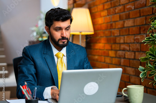 Asian businessman lost in thought while using laptop at desk in cozy workplace with brick wall