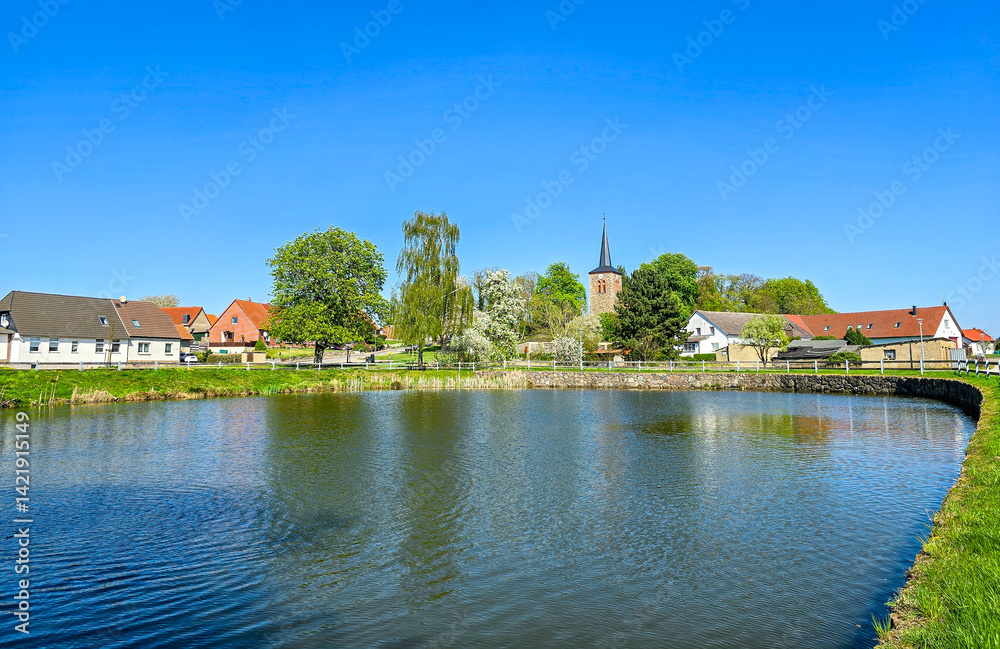 serene village with blossoming trees and a church spire reflects in a calm pond under a radiant blue sky, capturing a tranquil springtime atmosphere serene village with blossoming trees and a church spire reflects in a calm pond under a radiant blue sky, capturing a tranquil springtime atmosphere