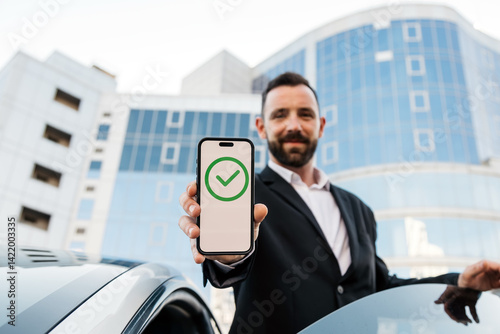 A businessman holds a phone next to his modern car with a sign of approved transfer or payment on the screen. The concept of confirmed, successful, completed or approved status.