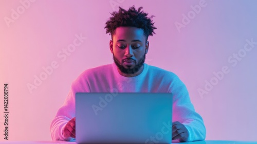 A young man with an afro sits at a table, intensely focused on his laptop under vibrant pink and blue lighting.