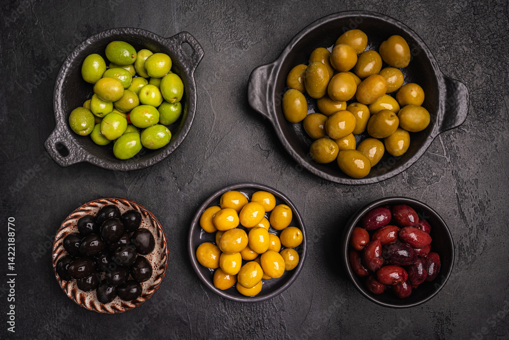 Green and black olives in a bowls and olive oil on dark background top view. Olive oil and variety of green and black olives in bowls with olive leaves all around taken from above.