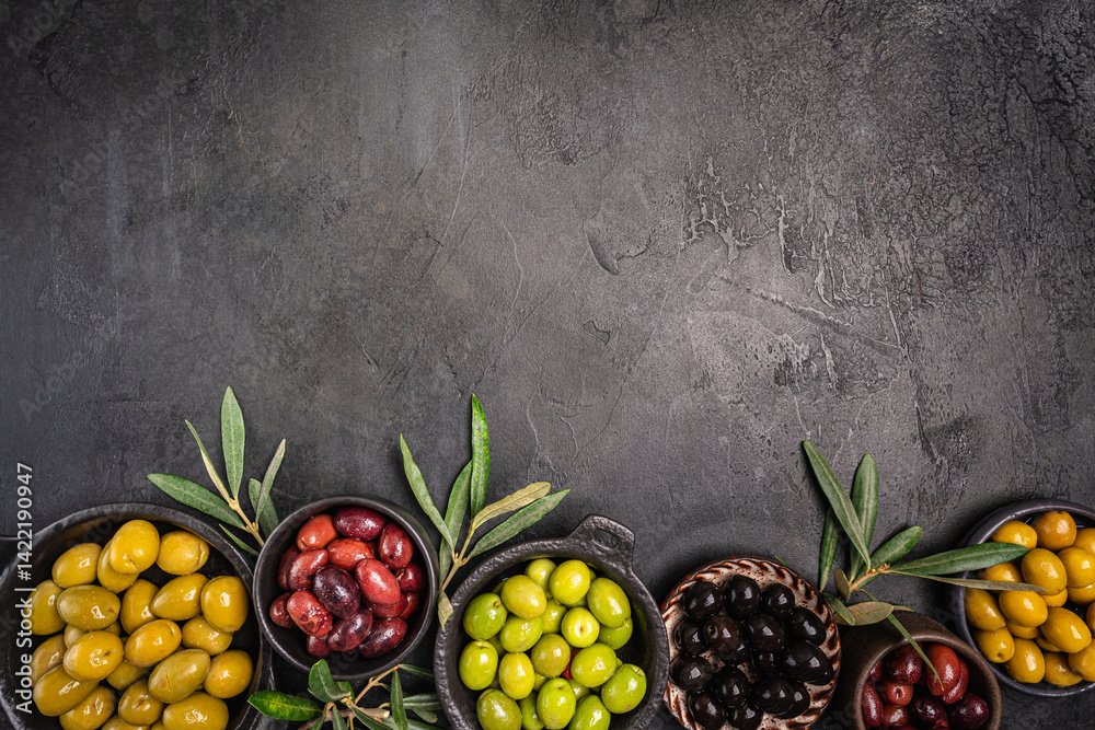 Green and black olives in a bowls and olive oil on dark background top view. Olive oil and variety of green and black olives in bowls with olive leaves all around taken from above.