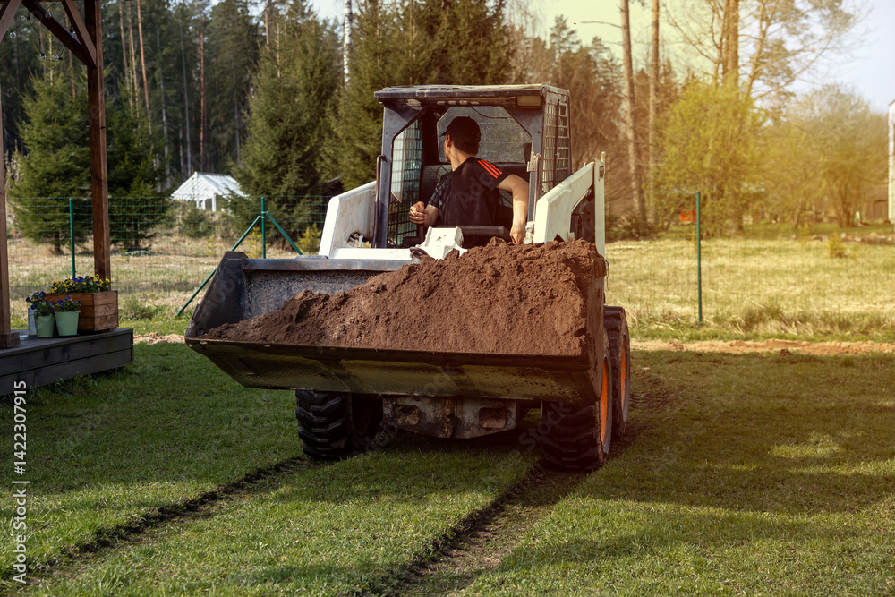 A man drives a skid steer loader carrying soil in an open grassy area. Tracks are visible on the grass, with a fence, forest, and small structure nearby.