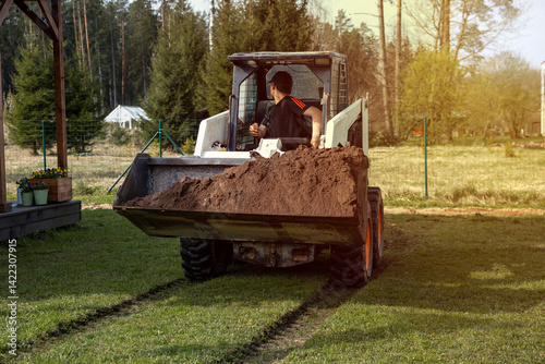 Obraz na płótnie A man drives a skid steer loader carrying soil in an open grassy area. Tracks are visible on the grass, with a fence, forest, and small structure nearby. w salonie