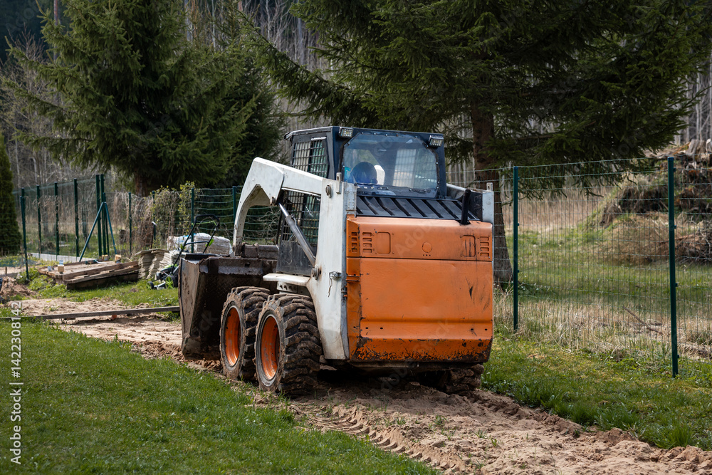 An orange and white skid steer loader rests on a dirt path with its bucket down. The area features tire tracks, disturbed soil, and tall evergreen trees.