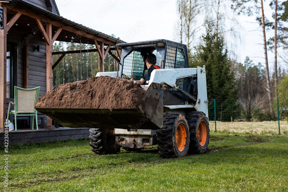 A skid steer loader with orange wheels moves soil on grass near a wooden house with a covered porch, chairs, string lights, trees, and a fenced area.